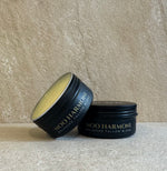 Two black metal tins of Moo Harmon Balanced Tallow Blend, one standing upright and one tilted on its side, showing the pale yellow tallow inside, set against a textured beige stone background.