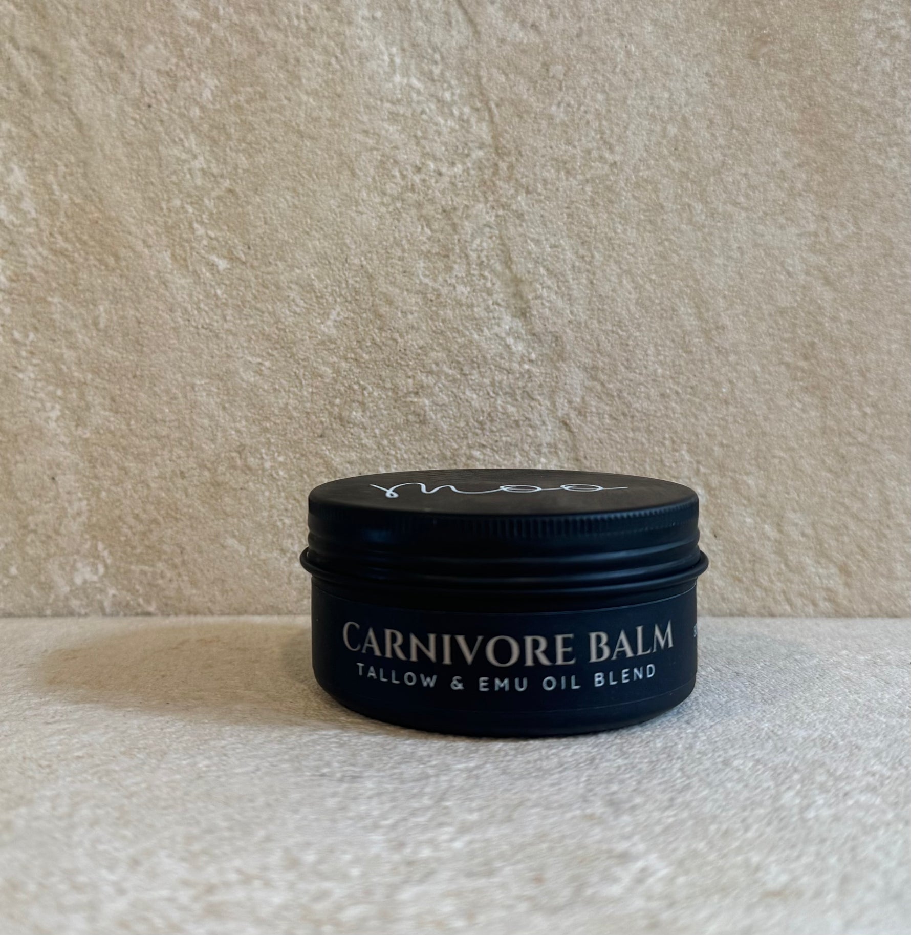 A small black metal tin of Carnivore Balm, a tallow and emu oil blend, set against a textured beige stone background.