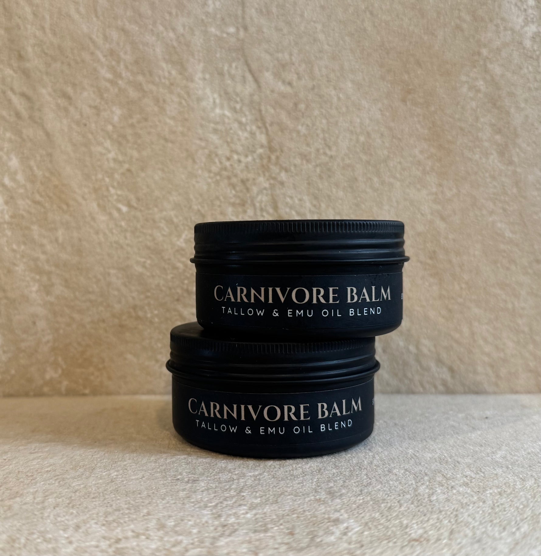 Two black metal jars of Carnivore Balm stacked on a beige stone surface against a textured beige wall. The labels read 'CARNIVORE BALM' and 'TALLOW & EMU OIL BLEND'.