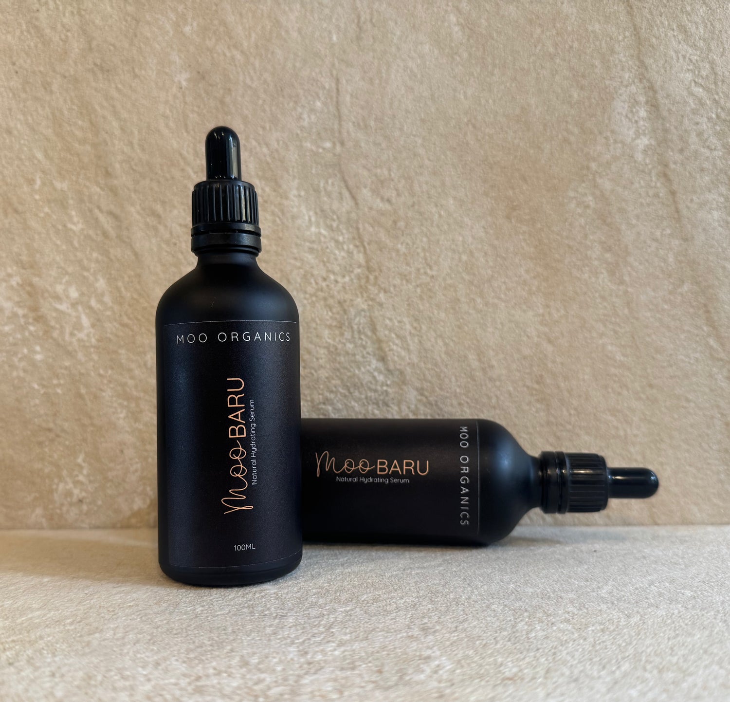 Two black dropper bottles of Moo Baru Natural Hydrating Serum by Moo Organics, one standing upright and the other lying down, set against a textured beige background.