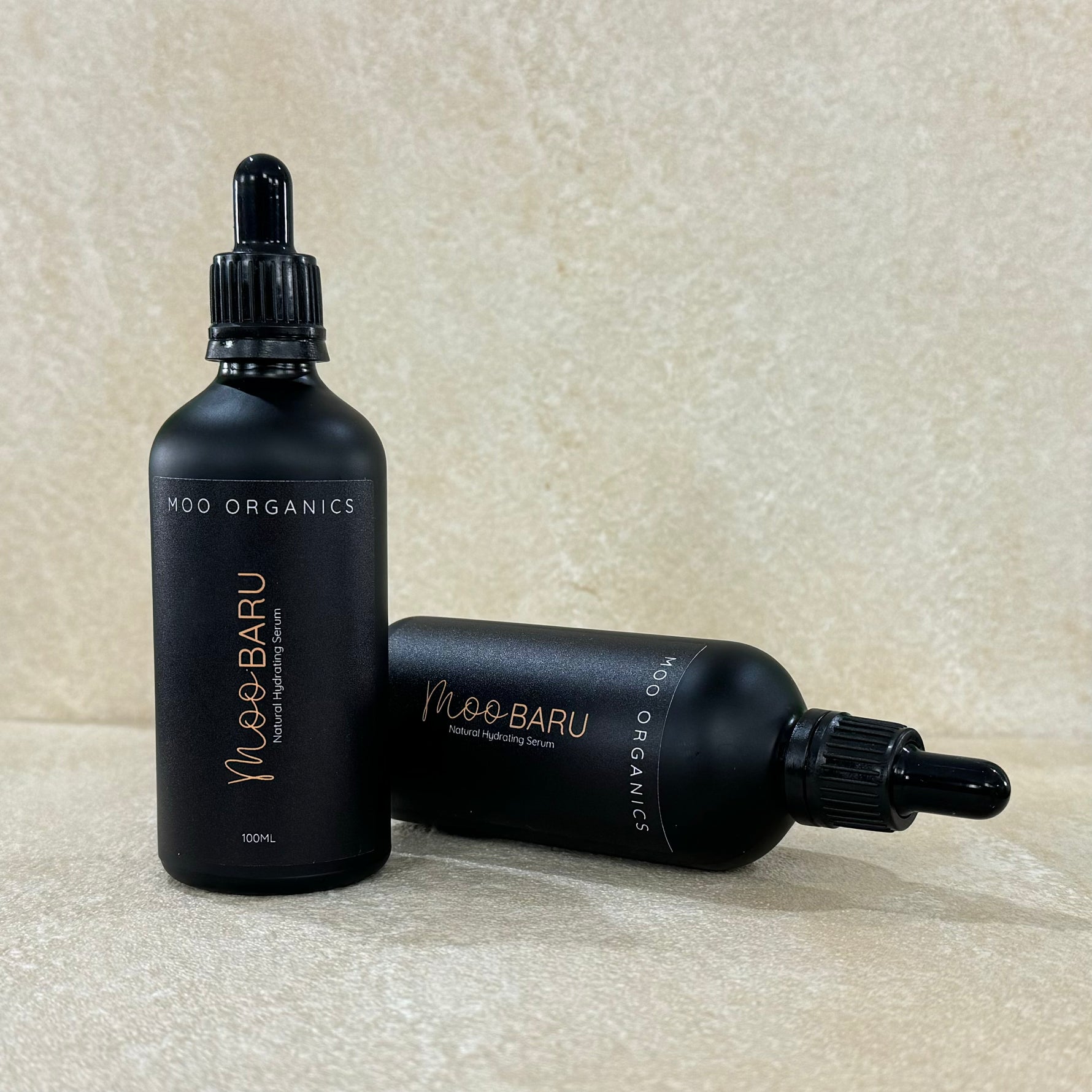 Two bottles of MOO BARU hydrating serum with black caps and labels, one standing upright and the other lying down, on a beige background.