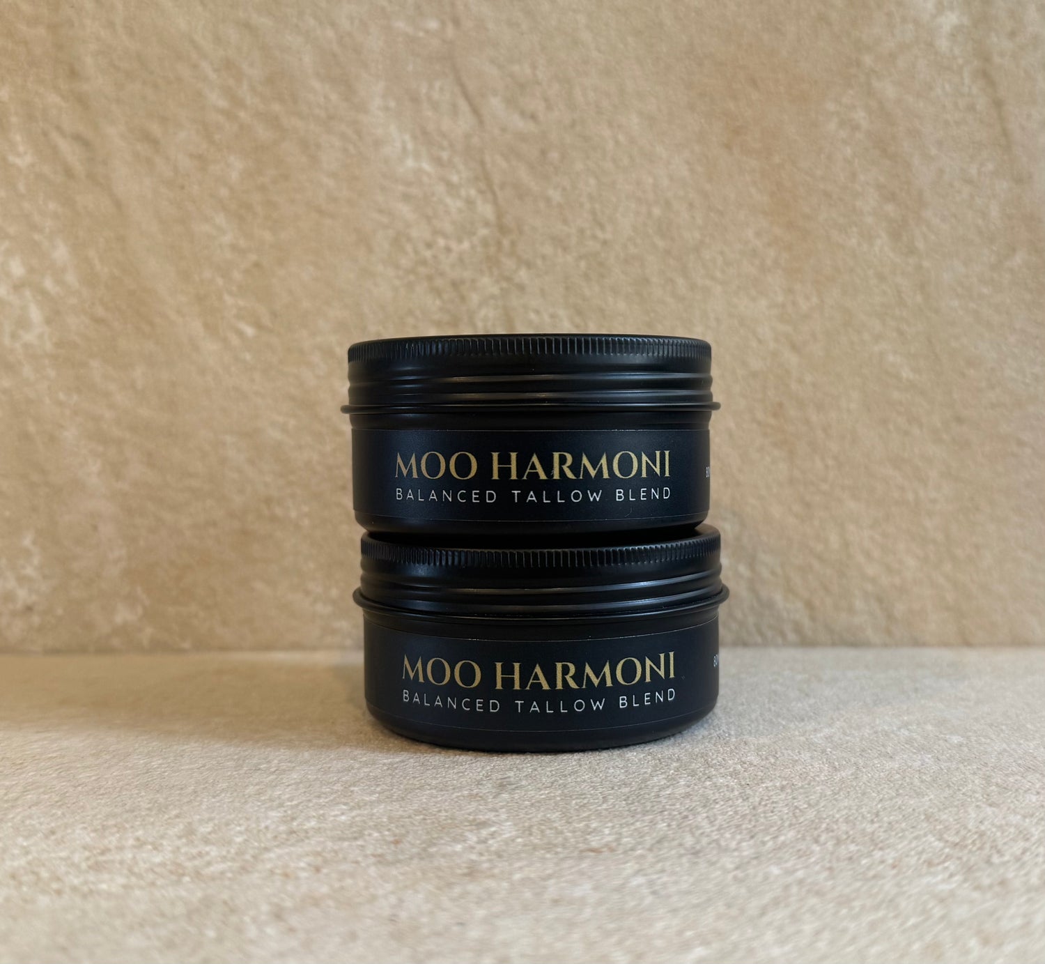 Two stacked black metal jars of Moo Harmoni Balanced Tallow Blend with gold lettering, set against a textured beige background.