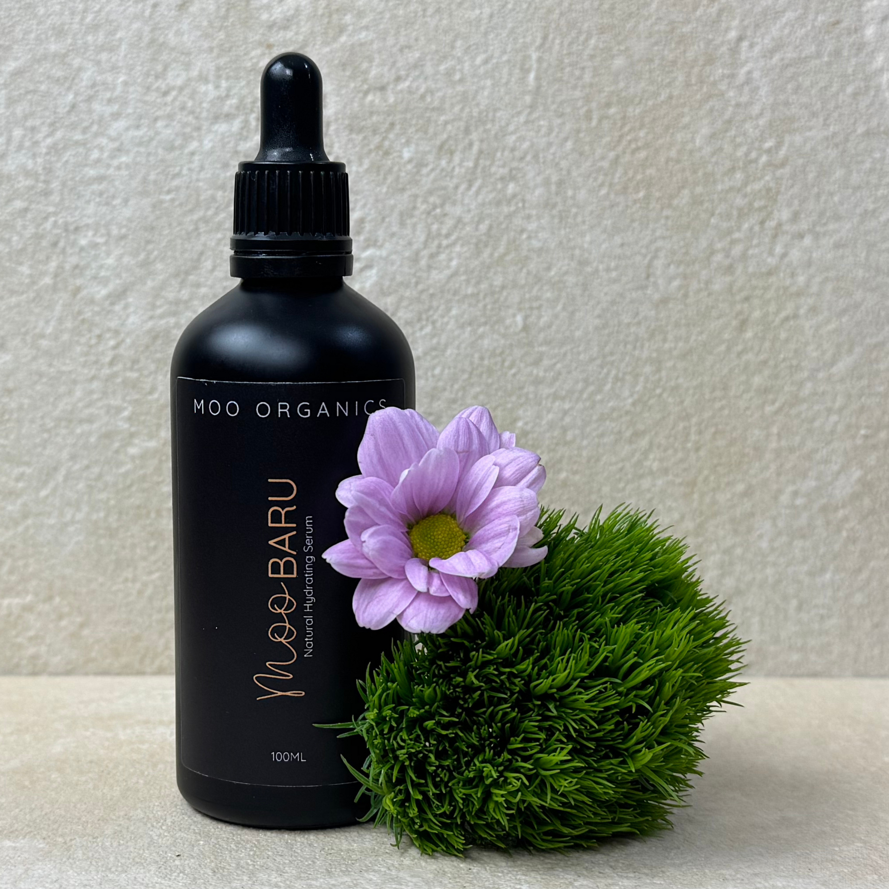 A black bottle of MOO ORGANIC MOO BARU Hydrating Serum with a dropper, displayed next to a purple flower and a small green mossy area, set against a beige background.