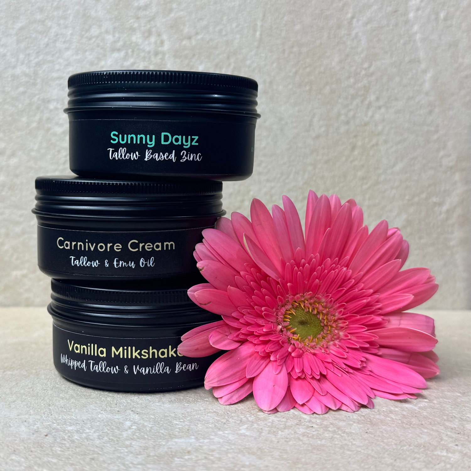 Three containers of Moo Organics skincare products labeled 'Carnivore Cream', 'Vanilla Milkshake', and 'Sunny Dayz' with a large pink flower in the foreground.