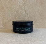 A black metal tin of Sunny Dayz Tallow Based Zinc Cream sits on a textured beige stone surface against a matching beige background.
