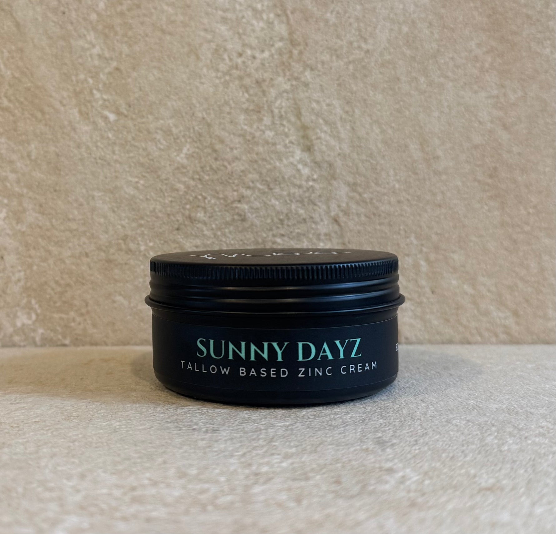 A black metal tin of Sunny Dayz Tallow Based Zinc Cream sits on a textured beige stone surface against a matching beige background.