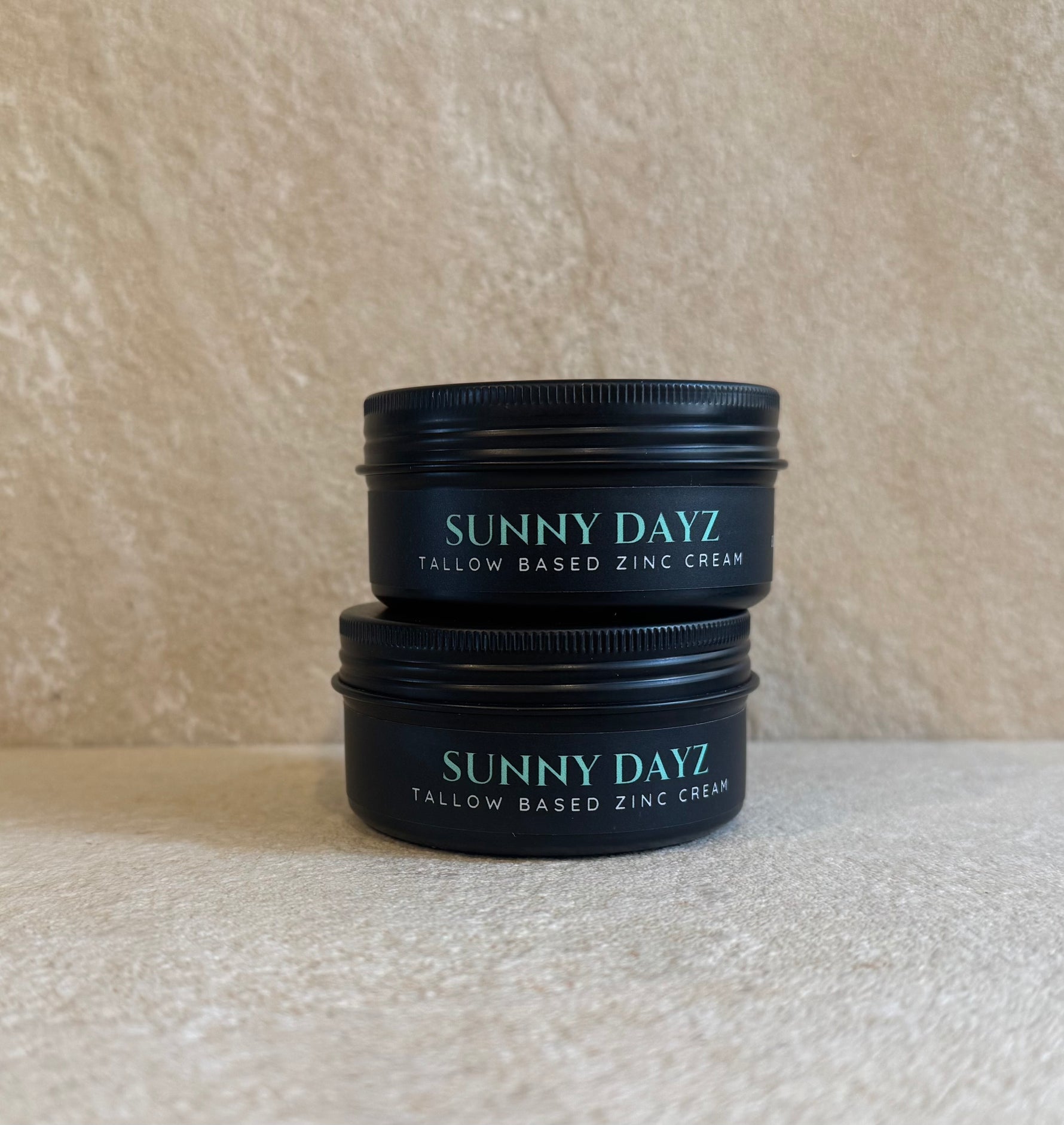 Two black metal tins of Sunny Dayz Tallow Based Zinc Cream stacked on a textured beige surface against a matching beige background.